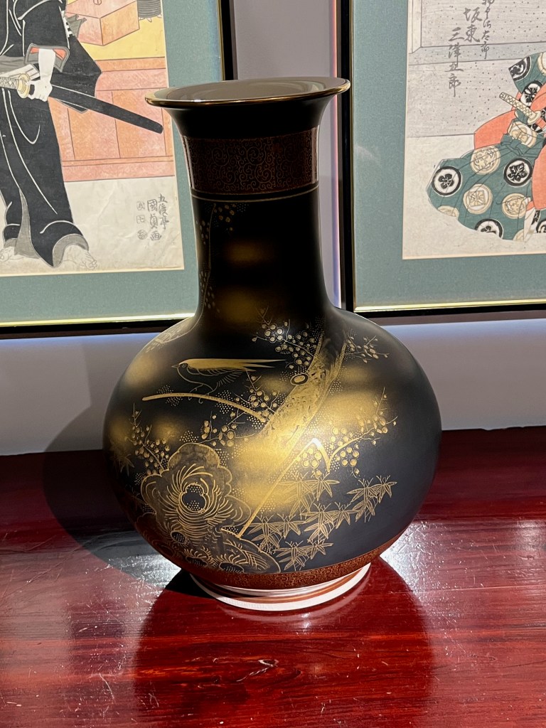 A large vase, consisting of a bulbous body with a large spout sticking up from the centre. The case is black, and the body is painted in gold to show birds in branches of plum, pine and bamboo trees.