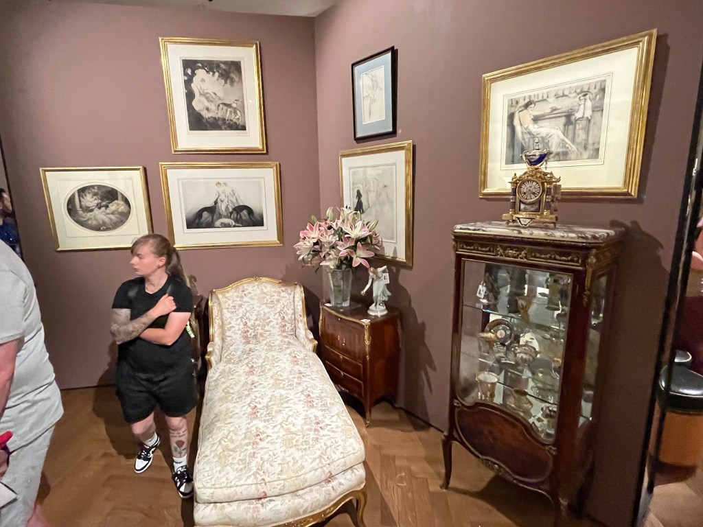 A day bed with a carved giltwood frame, and upholstered with a cream ground fabric decorated with flowering trees and landscape in light pink, purple, green and beige. There is a small wooden cabinet with a vase of flowers and a small statue of a woman near the top end of the bed, and a larger display case with a carriage clock on top at the opposite end. The furniture is set into a corner space, with 6 artworks spread across the walls.