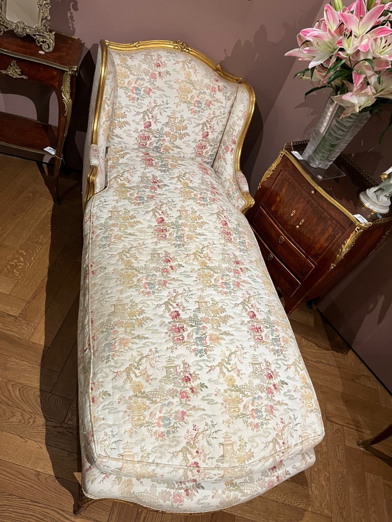 Viewed from the bottom end, a day bed with a carved giltwood frame, and upholstered with a cream ground fabric decorated with flowering trees and landscape in light pink, purple, green and beige.