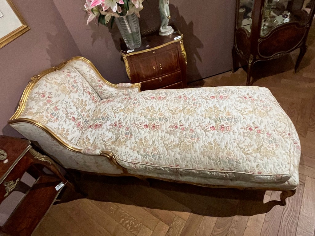 Viewed from the side, a day bed with a carved giltwood frame, and upholstered with a cream ground fabric decorated with flowering trees and landscape in light pink, purple, green and beige.