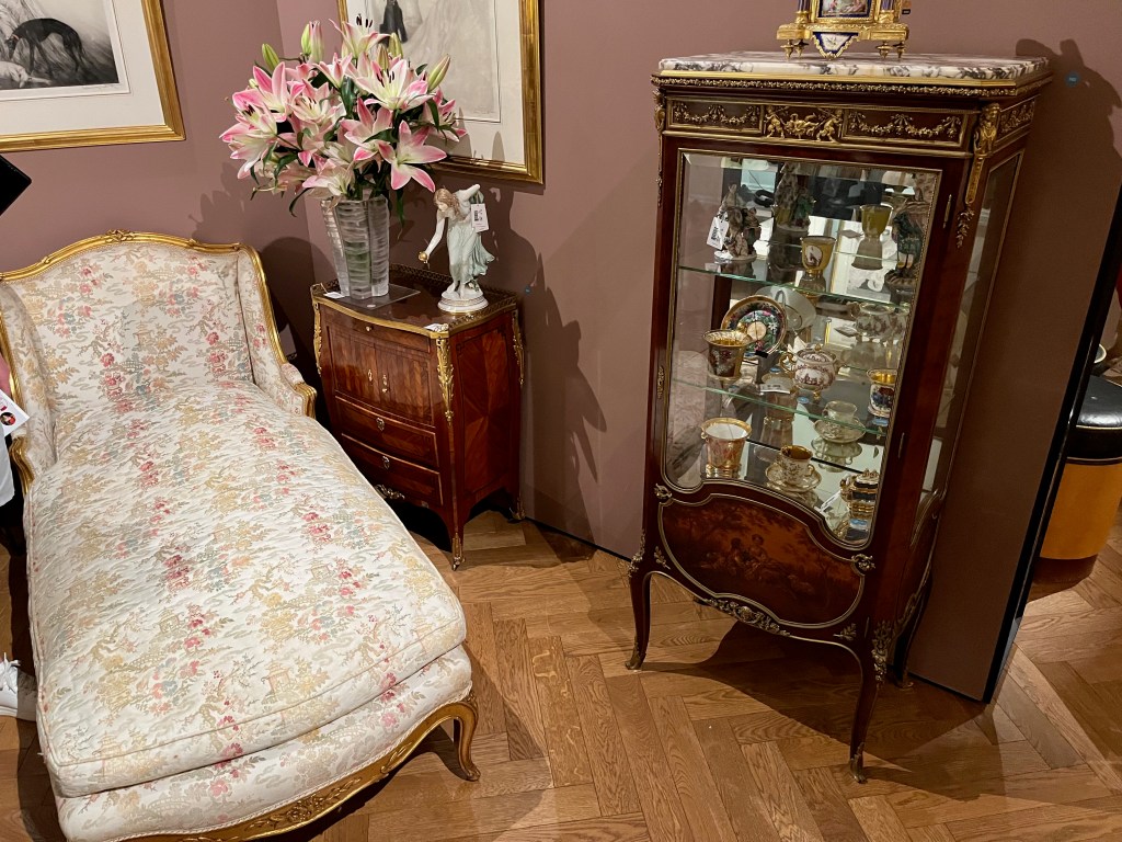 A day bed with a carved giltwood frame, and upholstered with a cream ground fabric decorated with flowering trees and landscape in light pink, purple, green and beige. To the right is a small cabinet, on which is a vase of flowers and a small porcelain statue of a woman leaning forward as she holds a round gold object in her hand, as if offering it to someone much smaller than her. Near the bottom end of the bed is a large display case with glass shelves holding colourful crockery and small statues.