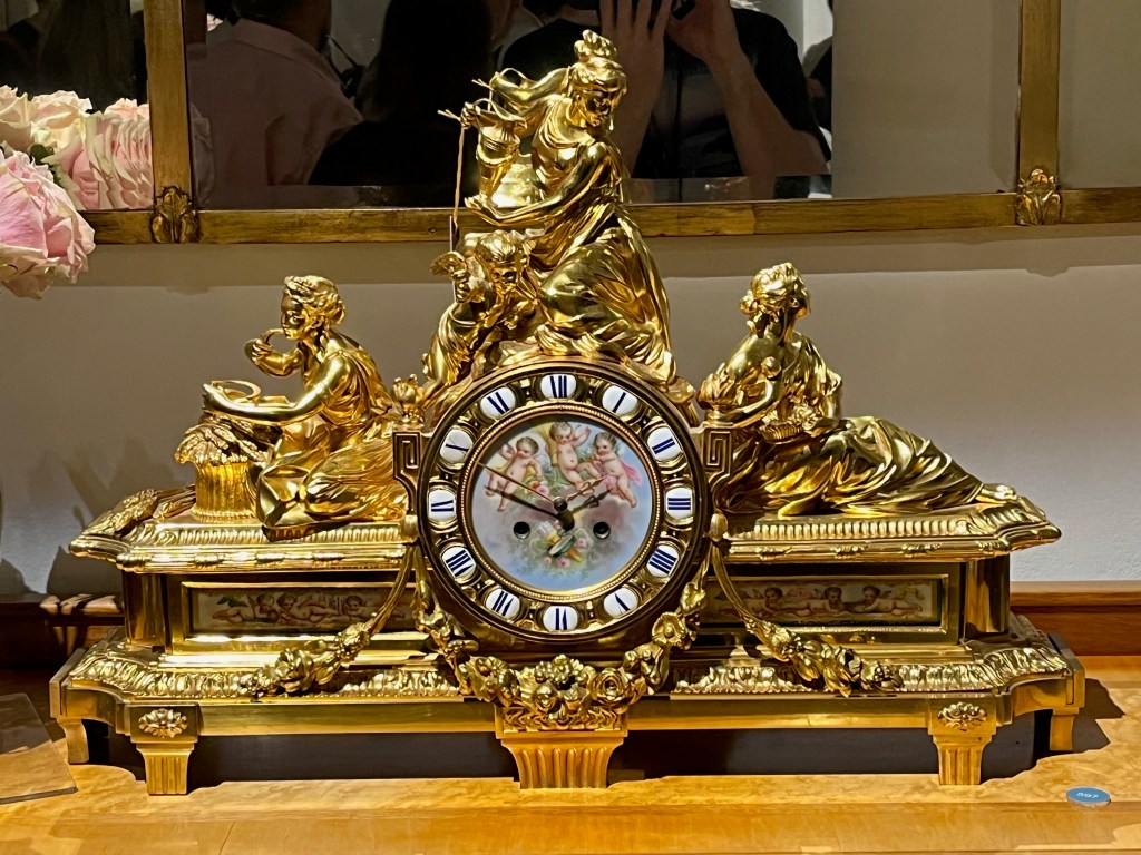 A very ornate golden mantel clock, with 2 classical features either side of the clock and one on top, while the background of the clock faces shows an image of three naked cherubs.