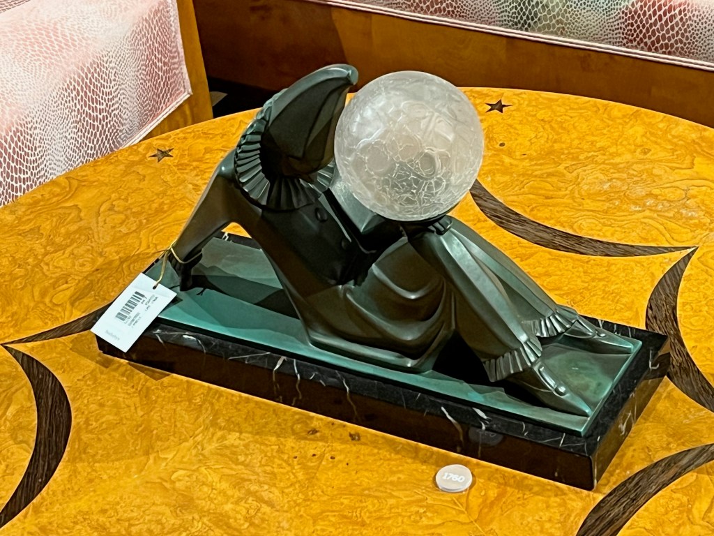 A table light. The black base resembles the figure of a person who is sat on the ground, with their legs bent in front of them so their feet are flat on the ground, while their arms are resting behind them to support them as they lean back. A large, ornately decorated glass ball, for the light, is balanced on the figure's bent knees.