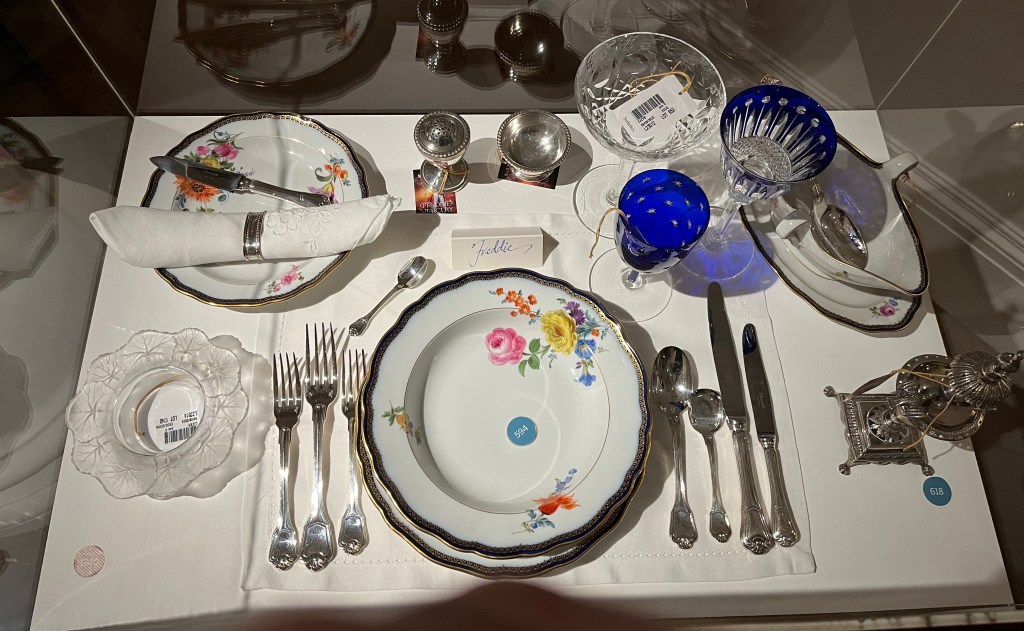 A dinner service featuring white plates painted with sprays of flowers within a blue and gilt-edged border. The plate has a wavy edge to it rather than being round. Also on the table are some cut glass Champagne glasses engraved with flowers.