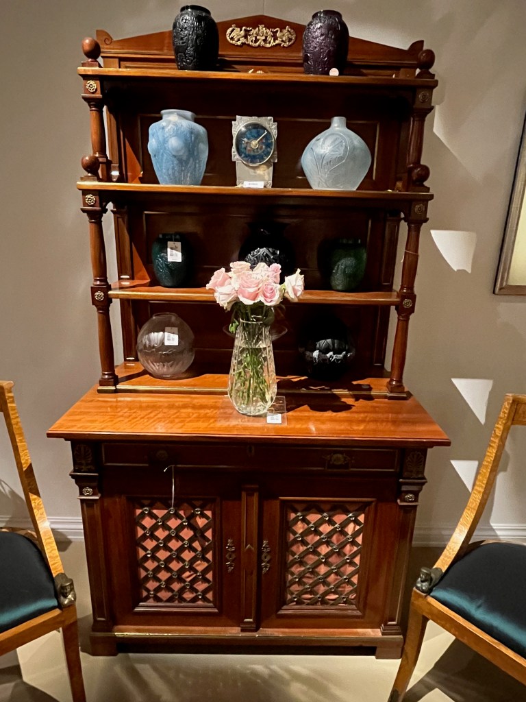 A tall chest with a cupboard beneath a counter at the bottom, 2 shelves above, and a ledge at the top. The cabinet is furnished with ornately sculptured vases, one of which has zebras on it, plus there's a mantel clock of the top shelf.