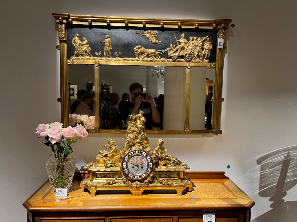A wide mirror with an upper section across its width featuring gold sculpted figures in a scene depicting a lion-drawn chariot, an angel and classically-dressed maidens. On top of a chest beneath it are flowers in a clear vase and an ornate golden mantel clock.