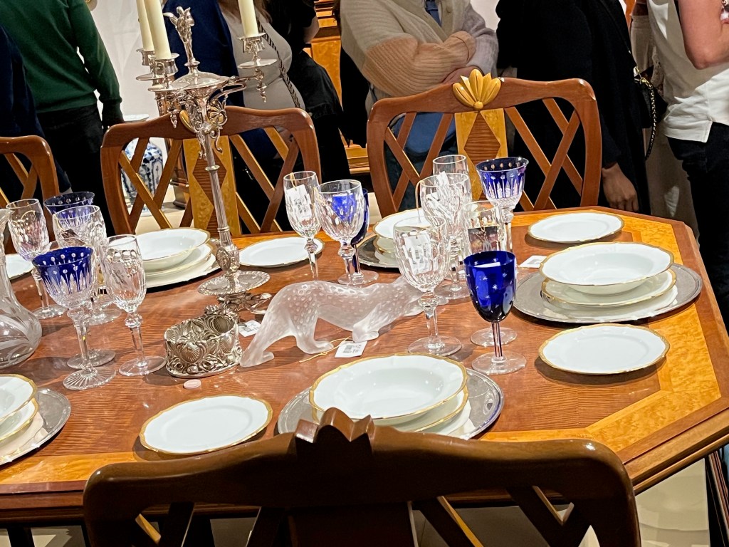 Right hand section of a large maple and mahogany dining table, filled with crockery and glasses, plus an ornate candle stick holder and other items. The most distinctive wine glasses and goblets have a deep blue colour with stars or Egyptian style elements.