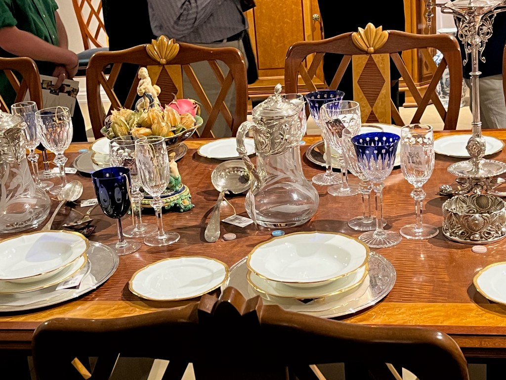 Middle section of a large maple and mahogany dining table, filled with crockery and glasses and other items. The most distinctive wine glasses and goblets have a deep blue colour with stars or Egyptian style elements.