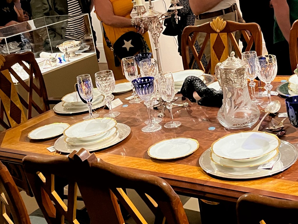 Left hand section of a large maple and mahogany dining table, filled with crockery and glasses, plus an ornate candle stick holder and other items. The most distinctive wine glasses and goblets have a deep blue colour with stars or Egyptian style elements.