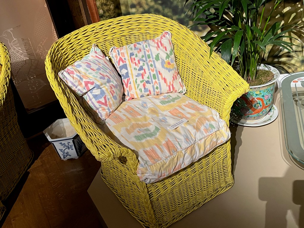 A wicker armchair in a yellowy-green colour, furnished with a seat cushion and two back cushions, which are white with a colourful pattern on them.