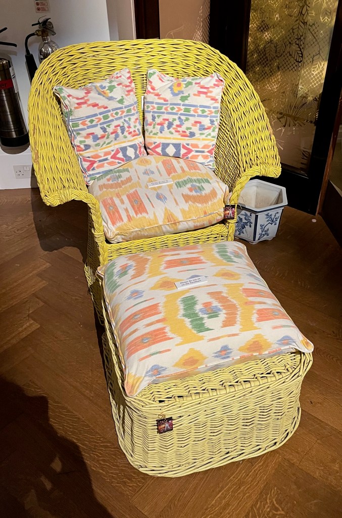 A wicker armchair in a yellowy-green colour, furnished with a seat cushion and two back cushions, which are white with a colourful pattern on them. In front of the chair is a matching foot stool, with a yellowy-green wicker base and a cushion ton the top.