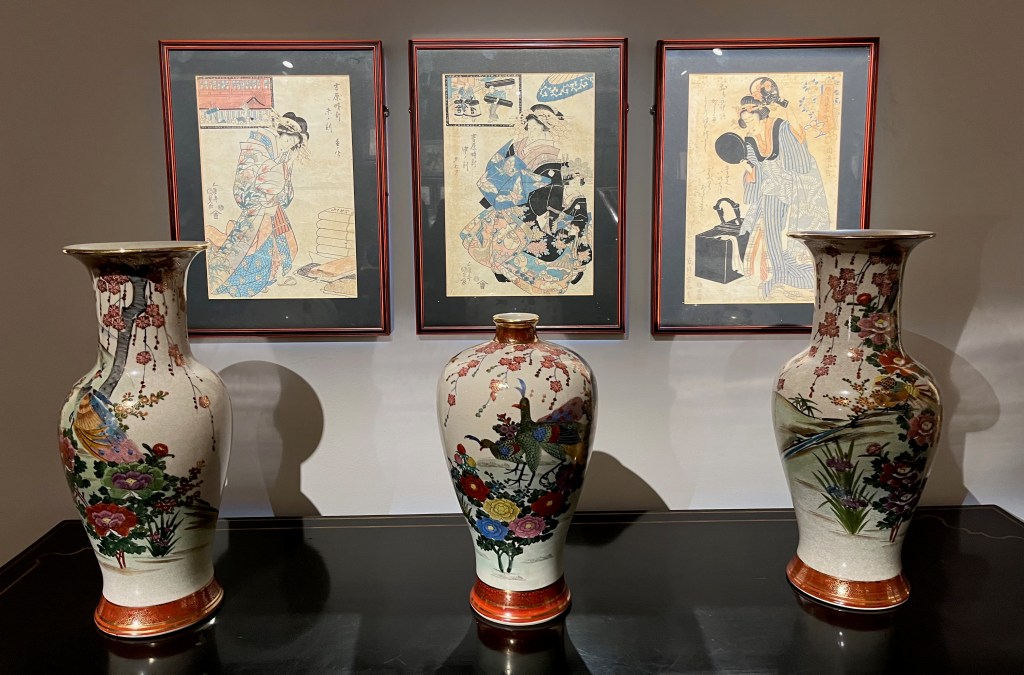 Three Japanese woodblock prints showing courtesan ladies in colourful kimonos and roads, on a wall behind three large white vases that are decorated with colourful outdoor scenes of birds and flowers.