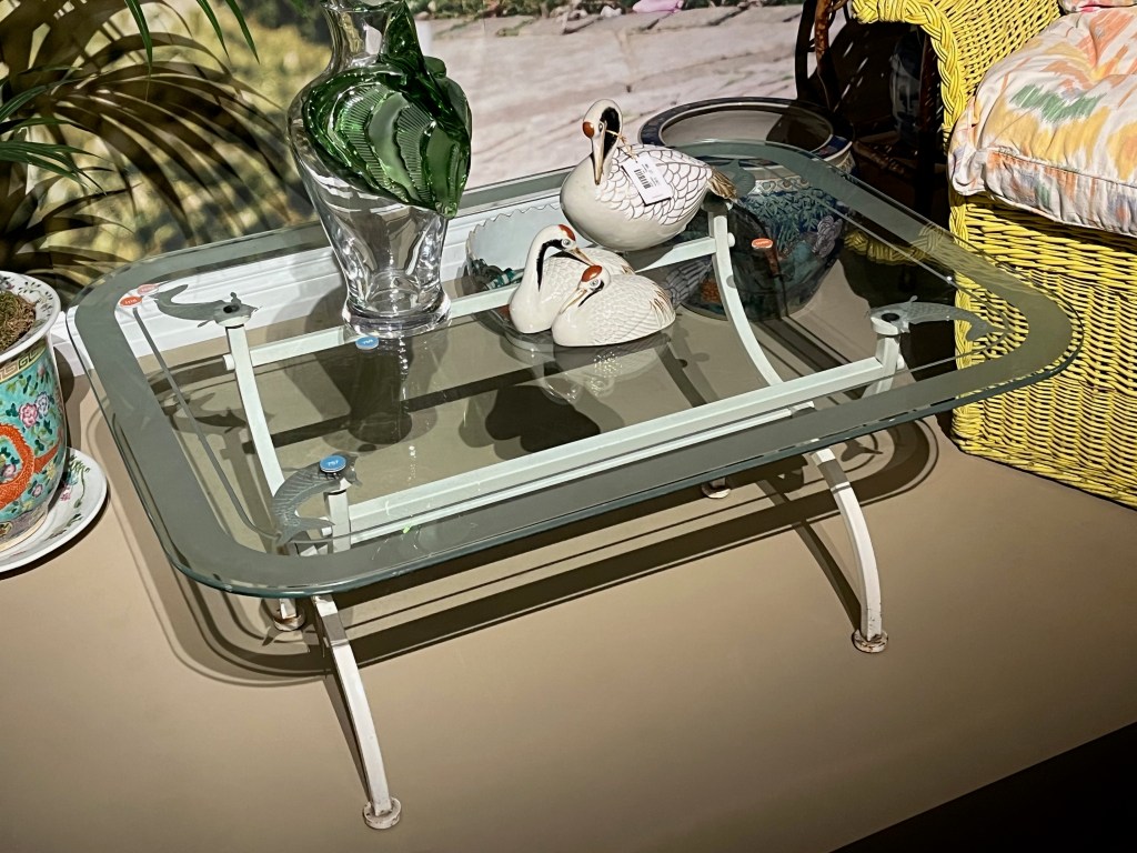 A table with a glass surface and metal frame, the corners of which have decorative carp fish on them. The table holds a large glass vase with a rippled design swirling around it, along with a few small white ornaments designed to look like swans.
