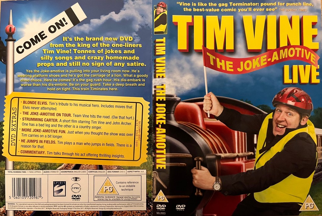 DVD cover spread for Tim Vine, Joke-Amotive. The front shows him grasping a rail on the side of a steam train engine with one hand, while using the other hand to hold up a small flag with the show's title. He's wearing a yellow high-vis jacket and a red helmet, and is smiling excitedly. The back cover shows a railway signal lifted that says Come On, but otherwise there is simply text about the show and extra features without any photos of Tim.