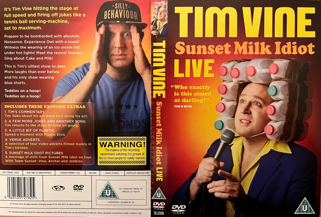 DVD cover spread for Tim Vine, Sunset Milk Idiot. The front shows him in front of a microphone, with 10 plastic milk bottles strapped around the left, top and right sides of his head with a lot of packing tape. The back shows him holding a hand up to each side of his face as he looks straight ahead, while wearing a black baseball cap with gold lettering saying Silly Behaviour.