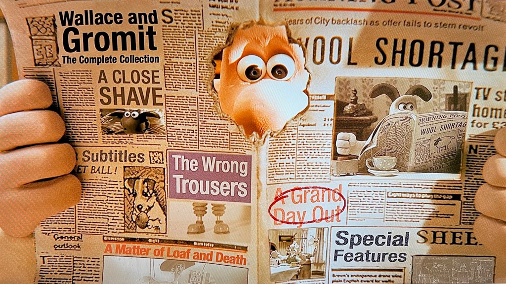 Main menu on the Wallace & Gromit Collection Blu-ray. It shows a spread open newspaper being held by Wallace, whose eyes we can see through a big hole in the top centre of the spread. The names of the short films, plus options for special features and subtitles, are spread around the newspaper to look like headlines. The headline image on the front cover of the paper, on the right of the spread, is a video showing clips from the films. In this screenshot it shows Gromit also reading a newspaper.