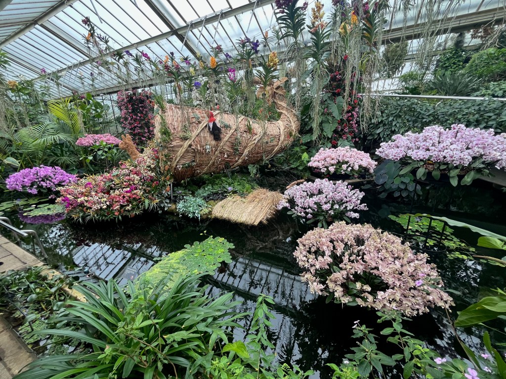 The large pond in Kew's Princess Of Wales Conservatory. In the centre, a large wicker-like tube is spilling out orchids in all sorts of colours, while a brown alpaca stands alongside it. Either side of that central display are arrangements of orchids in a range of colours, including white, pink and purple.