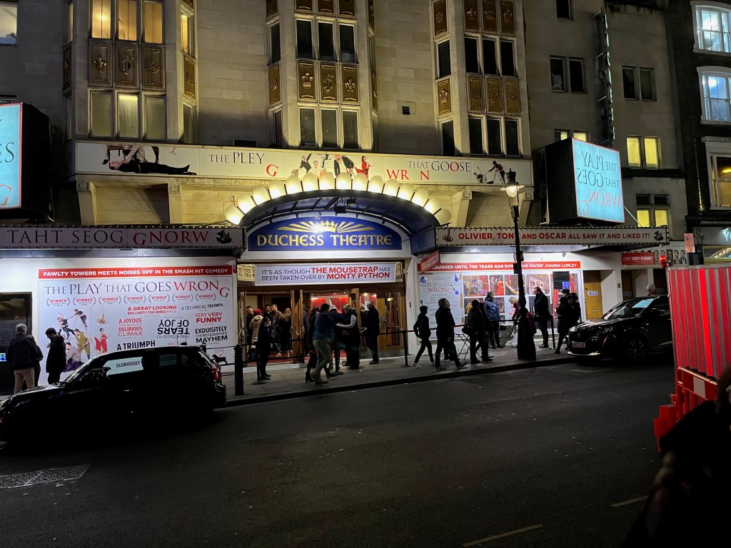 People entering the Duchess Theatre, which is lit up at night and has signage for The Play That Goes Wrong.