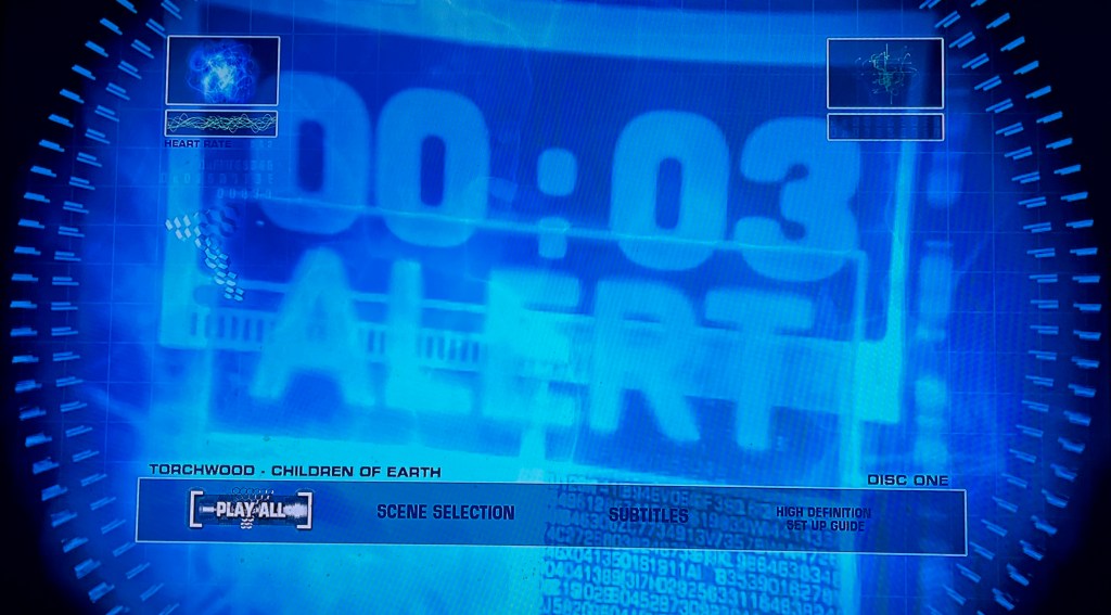Blu-ray menu for Torchwood, Children Of Earth, showing a blue-tinted image of a digital countdown clock that has 3 seconds remaining, with the word alert in big capital letters beneath it. The options along the bottom are Play All, Scene Selection, Subtitles and High Definition Set Up Guide.