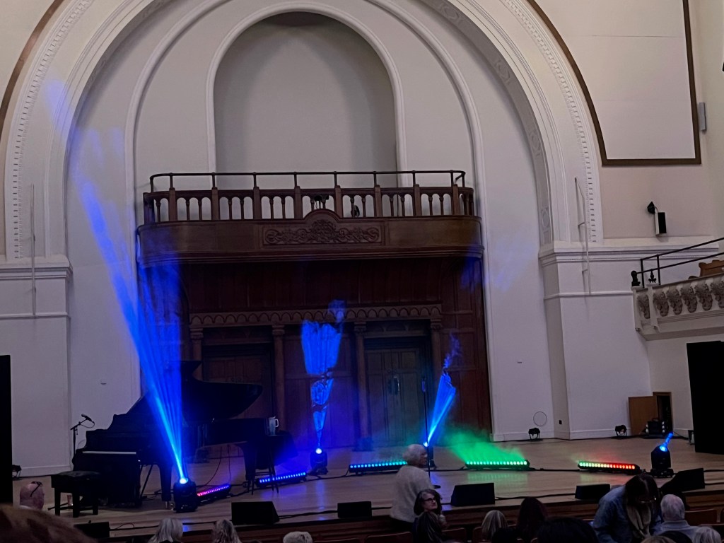 The stage in Cadogan Hall, in front of a massive arch in the white wall behind. The stage is a small wooden platform space with a semi-circle of lighting bars arranged on the ground to surround the performer, which are currently projecting blue and green colours up towards the ceiling. There is a piano on the far left of the stage area as well.