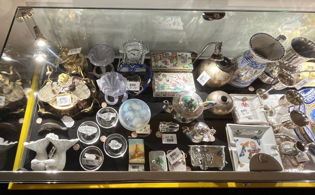 A selection of small objects in a glass case in the Freddie Mercury exhibition. Items include a crystal glass mantel clock, a brass inkstand, a paperweight in the shape of a heart, a model of a pair of doves, decorated boxes, ashtrays and silver teapots.
