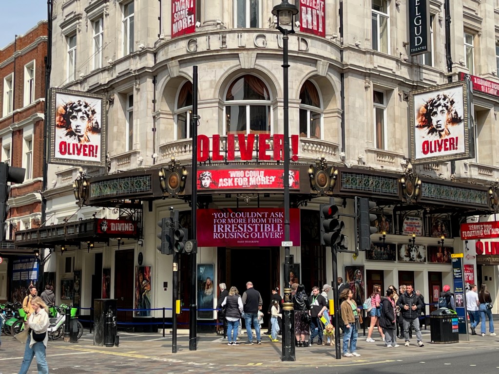 The entrance to the Gielgud Theatre, with signage for Oliver the musical. Attached to a balcony curving around the building, above the entrance, are big red letters spelling out Oliver, with an exclamation mark, and immediately below that is a red sign with white text that says Who could ask for more. A screen immediately above the ground floor entrance doors also has white text, which says You couldn't ask for more from this irresistible rousing Oliver.