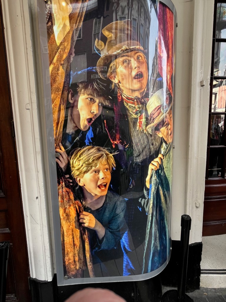 A poster for Oliver the musical, showing 4 members of Fagin's gang, including Oliver and the Artful Dodger, holding aside some curtains as they look outside with surprise and wonder.