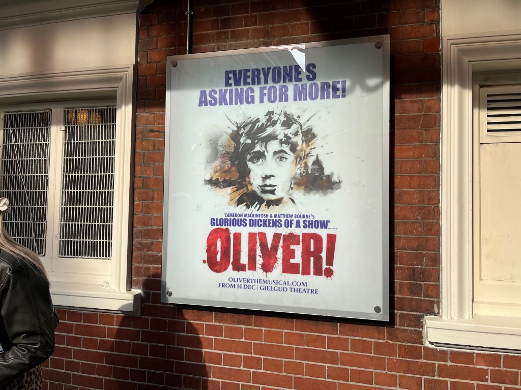Poster for Oliver, showing a painted image of a young boy's face, with the London skyline in the background, including St Paul's Cathedral. Blue text above him says Everybody's Asking For More. Just below the boy's face is more blue text saying Cameron Mackintosh and Matthew Bourne's glorious Dickens of a show. And immediately below that is the title Oliver, with an exclamation mark, in big red letters.