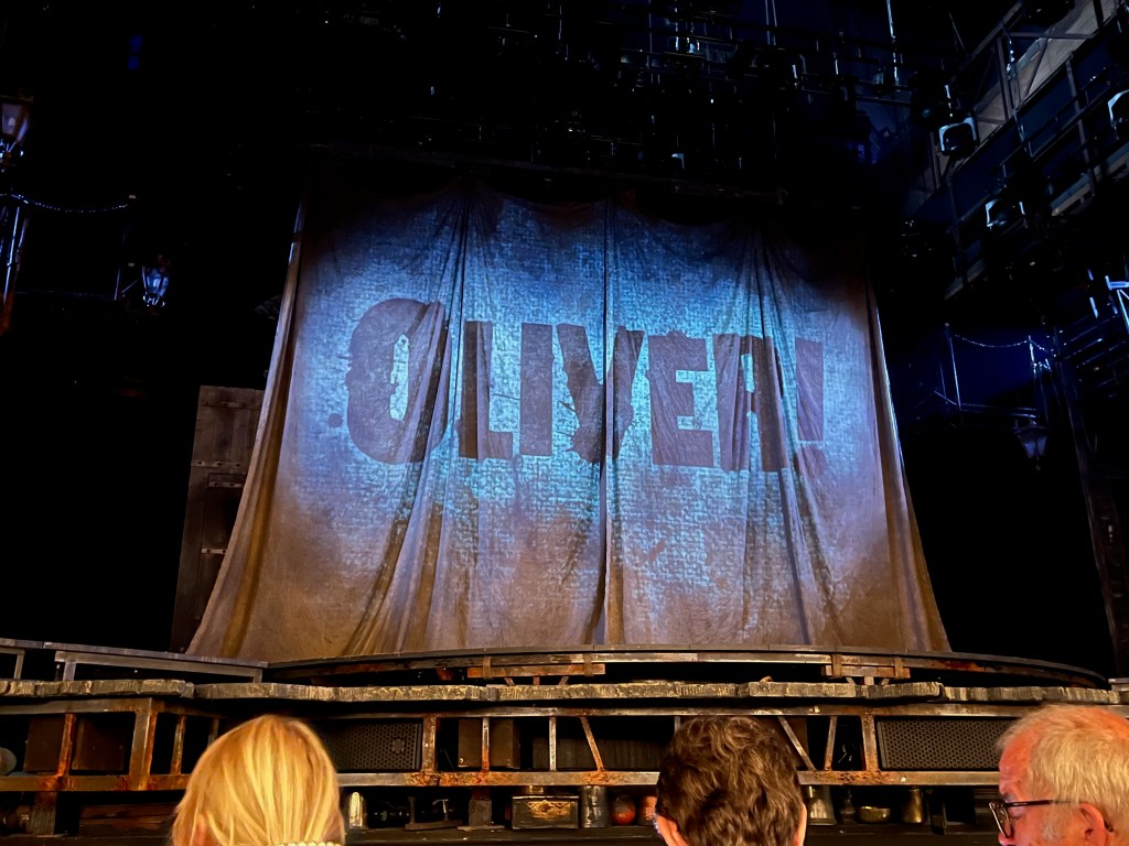 The stage at the Gielgud Theatre. The back half of the stage is concealed by a large dirty piece of cloth with the title Oliver displayed across it in large capital letters, with an exclamation mark at the end.