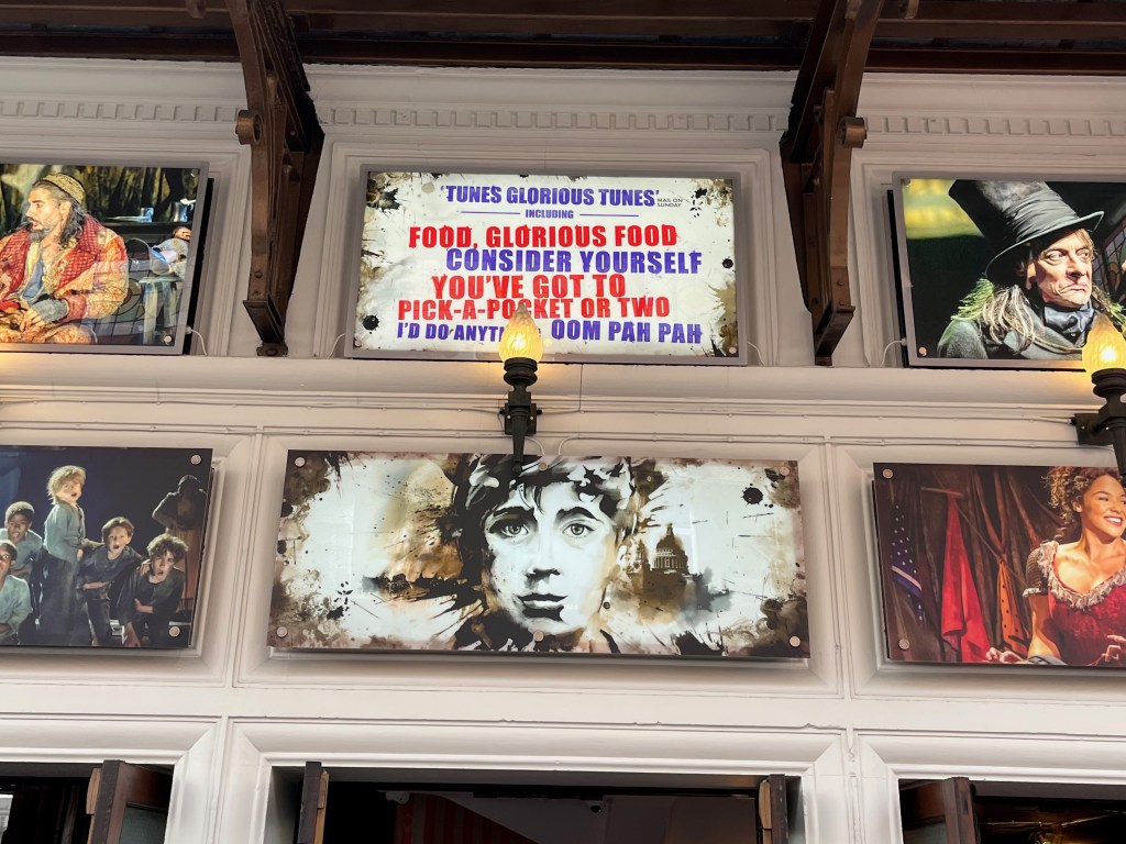 A white sign outside the Gielgud Theatre for Oliver the musical, with lines of blue and red text that say Tunes, Glorious Tunes, including Food, Glorious Food, Consider Yourself, You've Got To Pick-A-Pocket Or Two, I'd Do Anything and Oom Pah Pah.