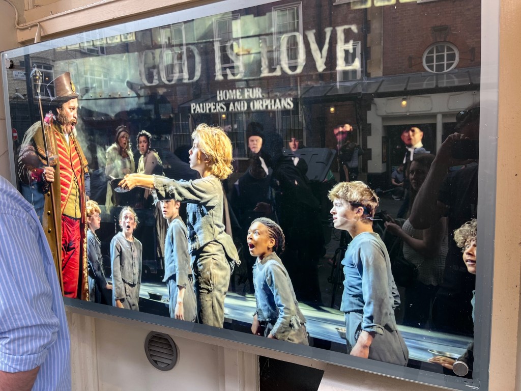 A poster for Oliver the musical, showing Mr Bumble looking incredulous in the workhouse as Oliver has the audacity to hold up his bowl to ask for more, while the other children look on in surprise and awe.