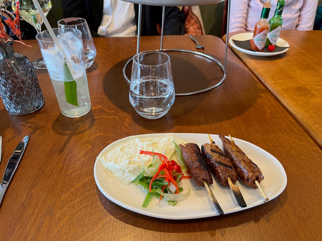 Lamb kofta, consisting of sausage-like cuts of lamb on small sticks, with whipped feta cheese to dip them into. Behind the dish is a glass of water, plus a Tropical Fizz cocktail, consisting of The Botanist gin, Noilly Prat vermouth, pineapple & fig leaf soda, plus it has a green leaf and a straw.