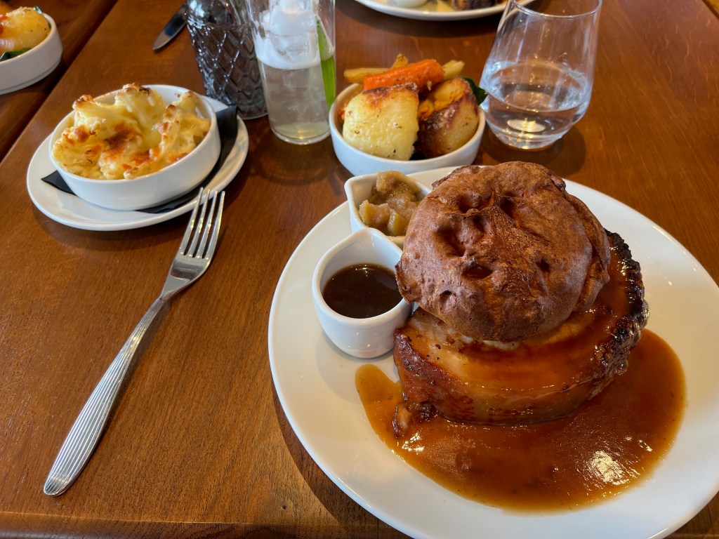A large round pork belly with a big Yorkshire pudding sitting on top like a hat. There is already some gravy on and around the pork belly, but there are small pots of gravy and apple sauce next to it. Behind the main dinner plate are 2 smaller bowls, one of which has cauliflower cheese, while the other has a couple of large duck fat roast potatoes and vegetables, including carrots.