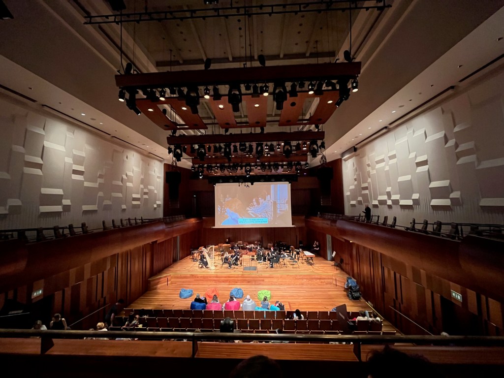 Milton Court Concert Hall. The National Open Youth Orchestra are setting up on stage, while a video about them plays on the screen on the back wall. As well as rows of seating, there are also some coloured beanbags right at the front that audience members can sit on.