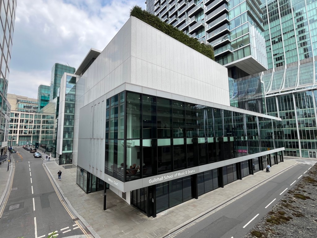 The large Milton Court building, with floor to ceiling windows on all three floors. A narrow grey wall separates the ground and first floors, on which is written Milton Court on one side of the building, and Guildhall School of Music and Drama on the adjacent side around the corner.