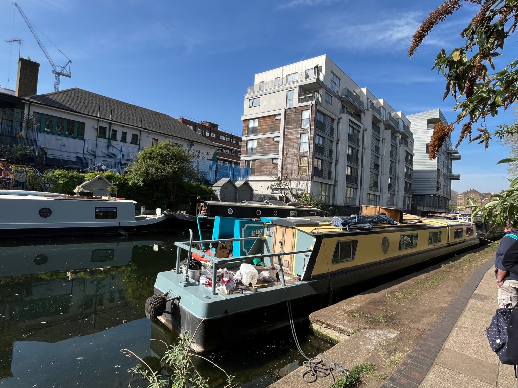 Long canal boats moored up next to the footpath on Regent's Canal, with tall blocks of flats on the opposite side of the canal.