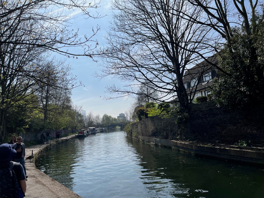 A view along Regent's Canal as it curves away into the distance, with trees on each side and boats moored up by the footpath on the left.