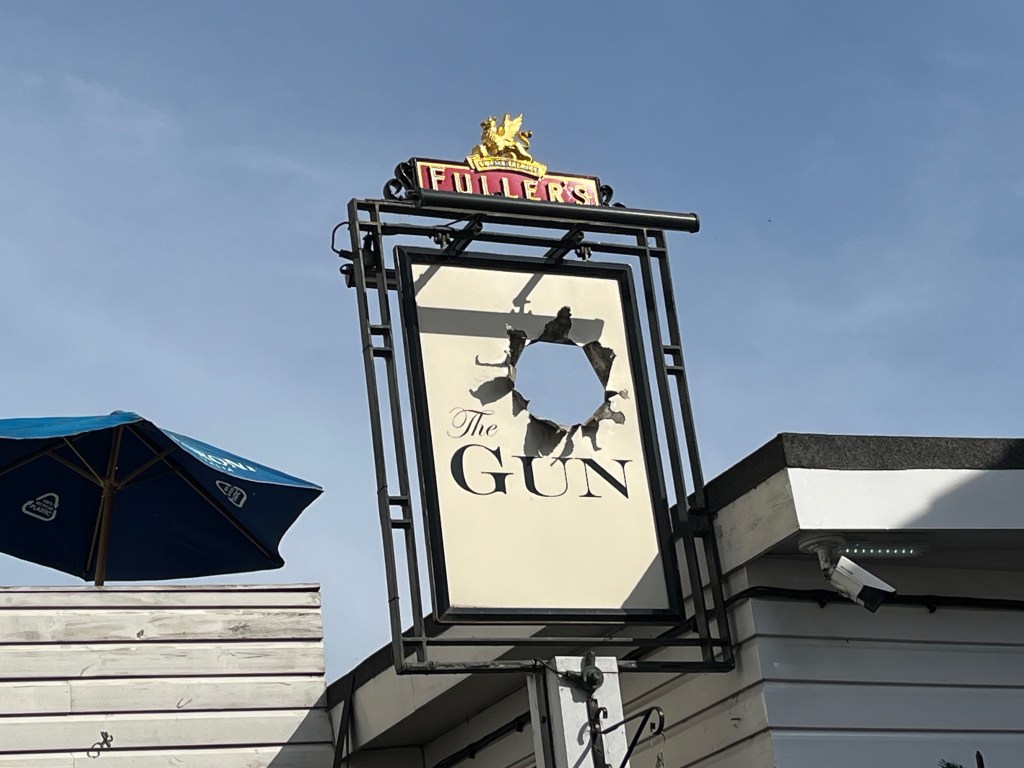 Sign for The Gun pub. It's in a portrait orientation, and is white within a black frame, with black text saying The Gun. Just above the name, taking up most of the top right corner, is a large hole in the sign with torn edges, to make it look as if something has been fired through it.