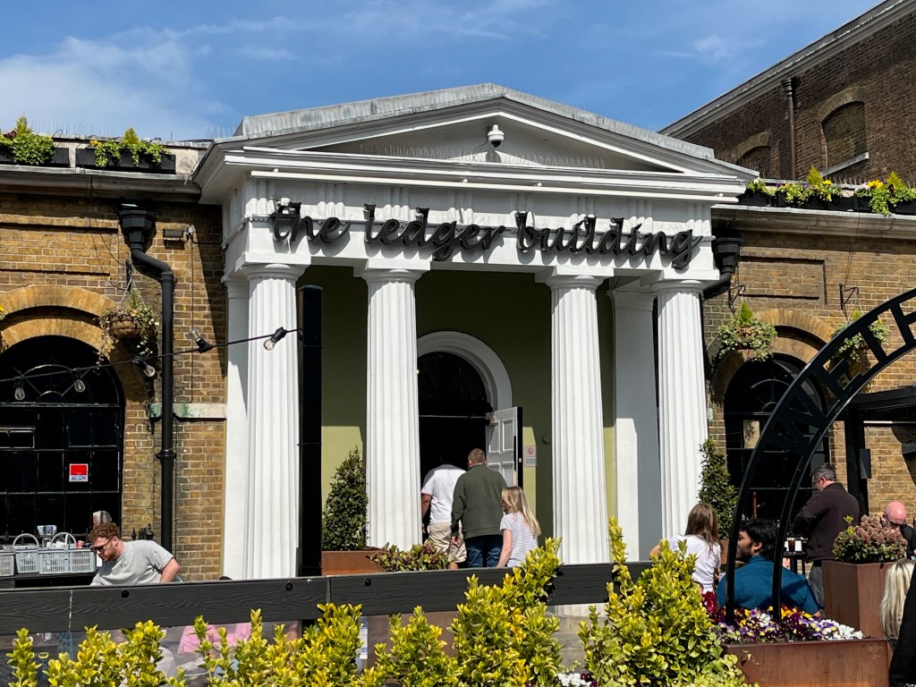 The entrance to The Ledger Building, a Wetherspoon pub. To get to the door you pass through a large white  portico with 4 pillars and a triangular roof. On the wall between the top of the pillars and the roof is large black text in a lowercase handwriting style that says The Ledger Building.