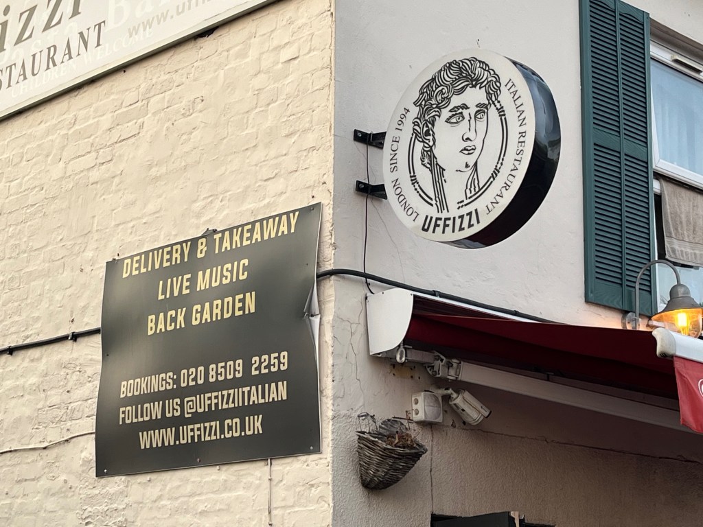 A round sign for the Uffizzi restaurant sticking out from the front of the building, It has a lady's face in the centre, while text around the edge says Italian restaurant Uffizzi London since 1994. Meanwhile a black sign with gold text on the side of the building says delivery and takeaway, live music, back garden. Bookings 020 8509 2259. Follow us at UffizziItalian. www.uffizzi.co.uk
