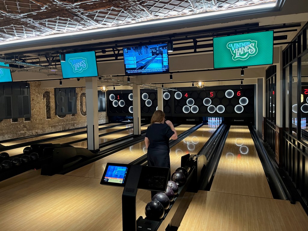 A row of 6 ten-pin bowling lanes at Vintners Lanes, with a screen above each one. At the far end, on the wall above the openings for the skittles, are circular lights arranged in a random configuration, and red lane numbers. Emily can be seen preparing to bowl down one of the lanes.