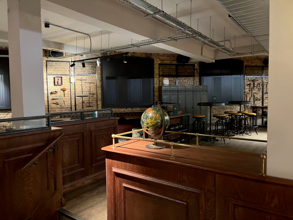 Part of the seating and dining area at Vintners Lanes, including stools arranged around circular tables and a set of 6 tall grey storage lockers. A narrow wooden counter with a railing around the edge at the entrance to this area has a globe on a stand.