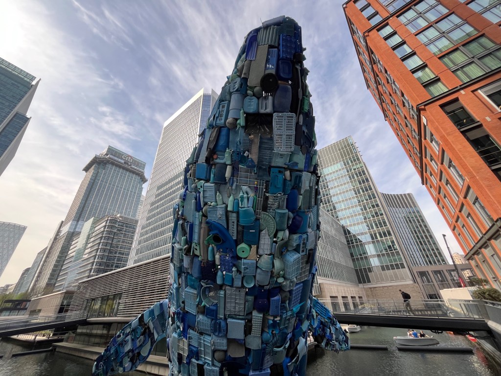 A huge 11-metre tall model of a blue whale standing tall out of the water in Canary Wharf. It is made completely out of plastic litter, including bottles, boxes and more.