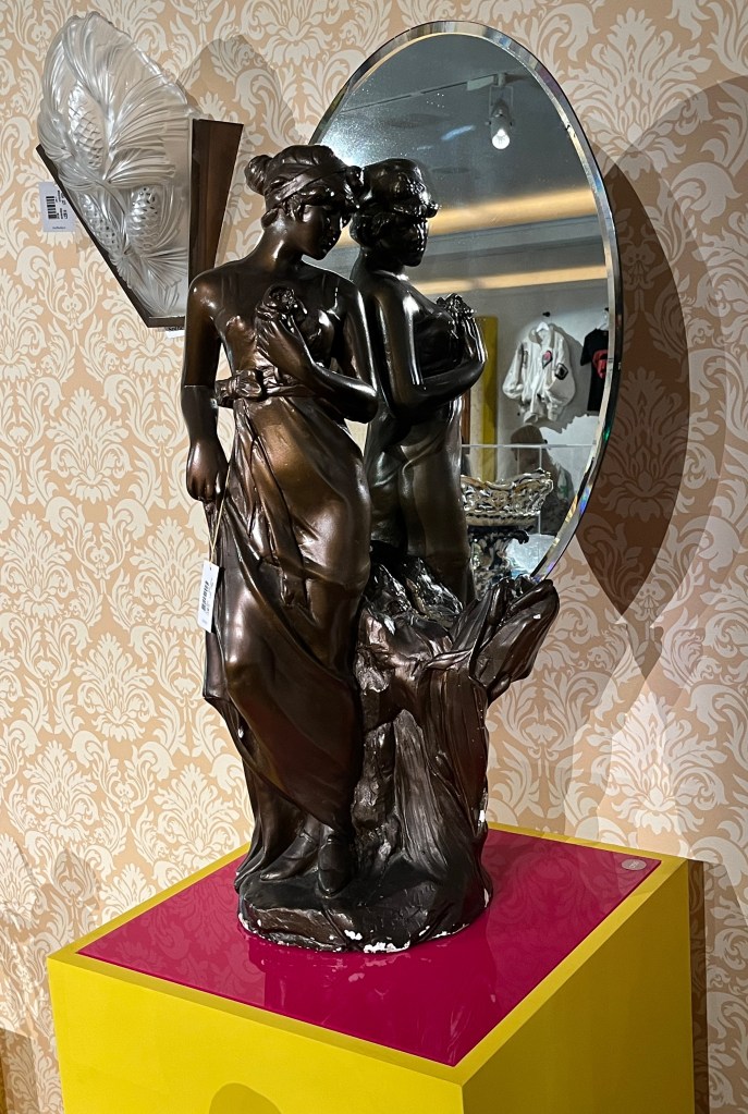 An oval mirror being held by a bronze sculpture of a maiden holding a rose next to a tree stump.