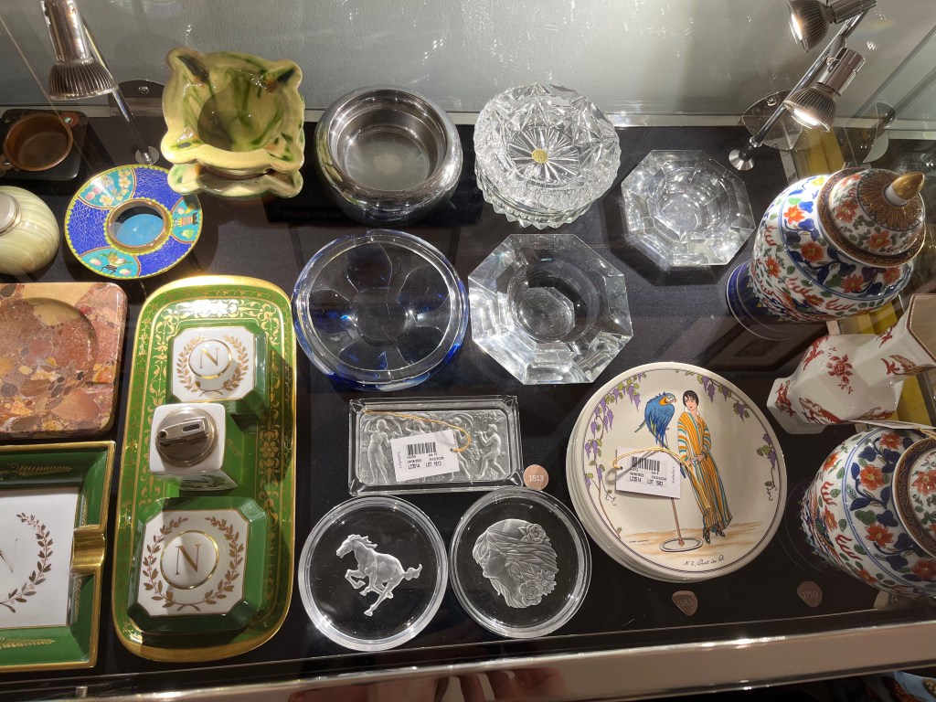 Ten glass ashtrays in a variety of designs, plus a stack of plates and a set of three vases that have colourful designs on them.