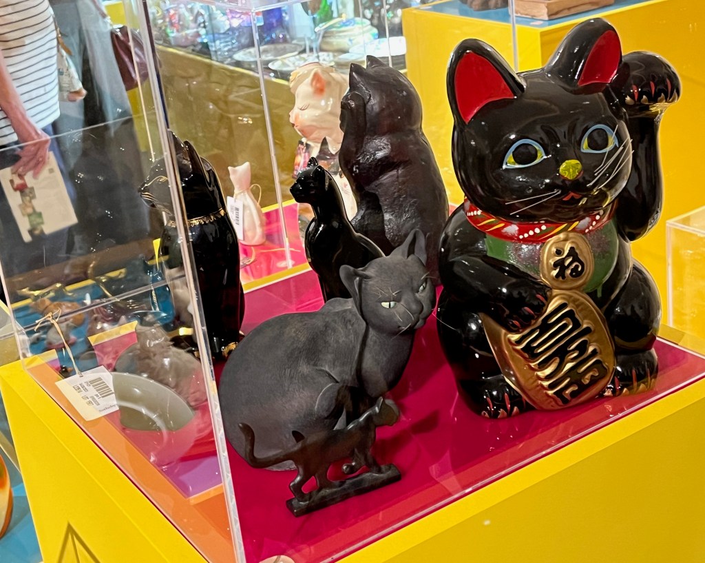 Glass case containing ornaments of black cats in varying designs and sizes. The largest has red pointed ears, blue edging to its eyes, white whiskers, a small yellow nose, an even smaller red mark for its mouth, and a red scarf with colourful patterning around its neck, and it's hold a round gold object in front of it with one of its paws.