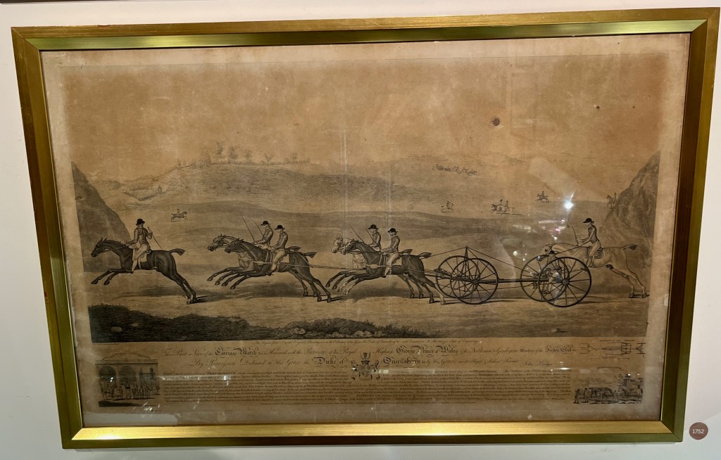 The painting Carriage Match at Newmarket by John Bodger. It shows 5 people on horseback, arranged as one leading two pairs, pulling a simple vehicle that's just a narrow platform with 4 big wheels, while one more person on horseback follows behind. They are moving at speed through a landscape with hills in the background.