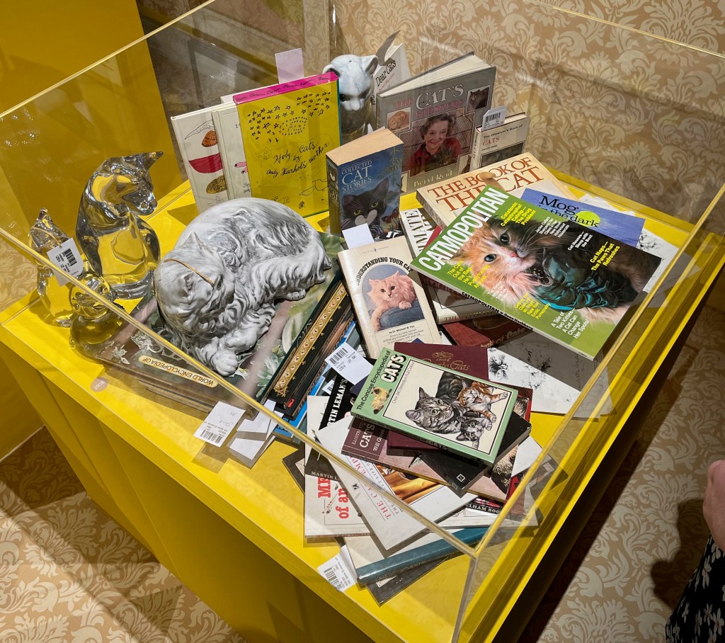 A glass case containing three glass seated figures of cats, two porcelain seated models of cats, and a collection of 29 books about cats.