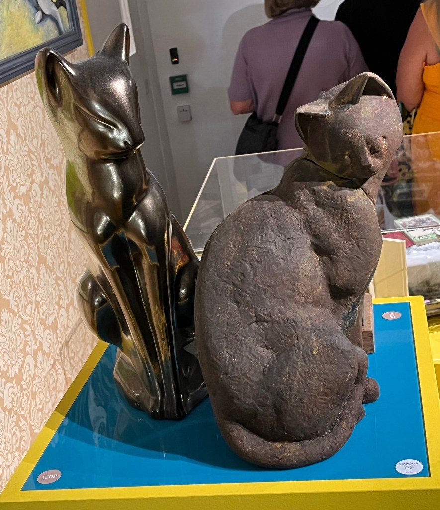2 ornaments of cats, one of which is sitting with its tail curled around its body, as it looks back over its shoulder at us. The other cat figure is viewed from the back