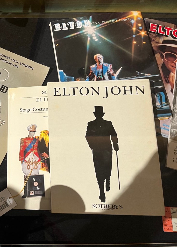 Items relating to Elton John. There are 2 Sotheby's catalogues, one of which has a silhouetted image of Elton walking away with a top hat and walking stick on the cover. And behind those is a book called Elton - It's a Little Bit Funny, which shows him performing on stage on the cover.